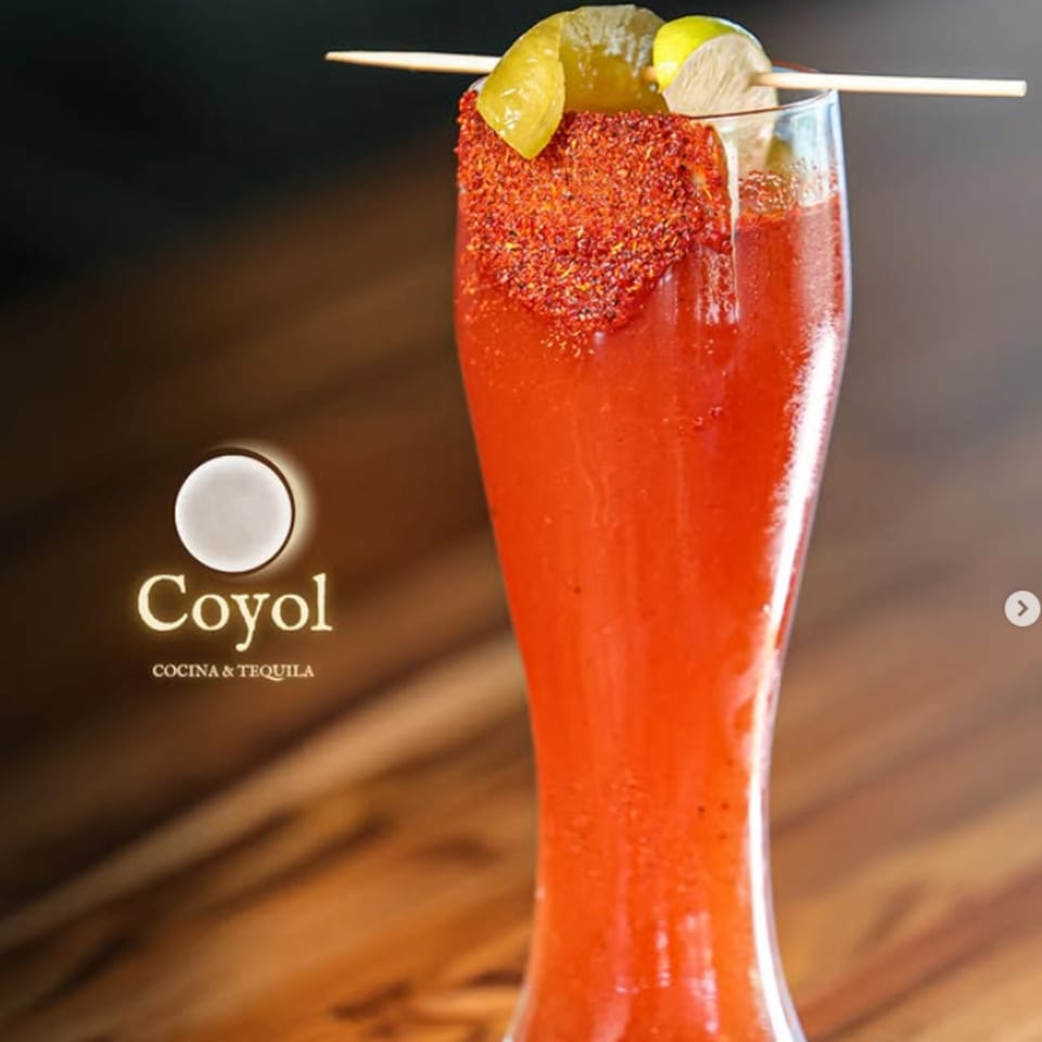 Tall red cocktail with a chili-salt rim, lime wedges, and a wooden skewer at Coyote Cocina & Tequila.