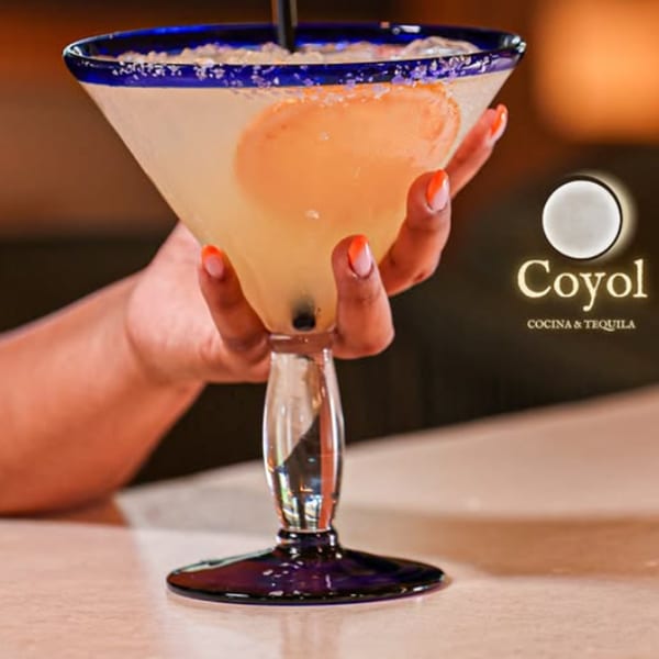 Hand holds a blue-rimmed margarita glass filled with a pale orange cocktail beside a Coyol Cocina & Tequila sign.