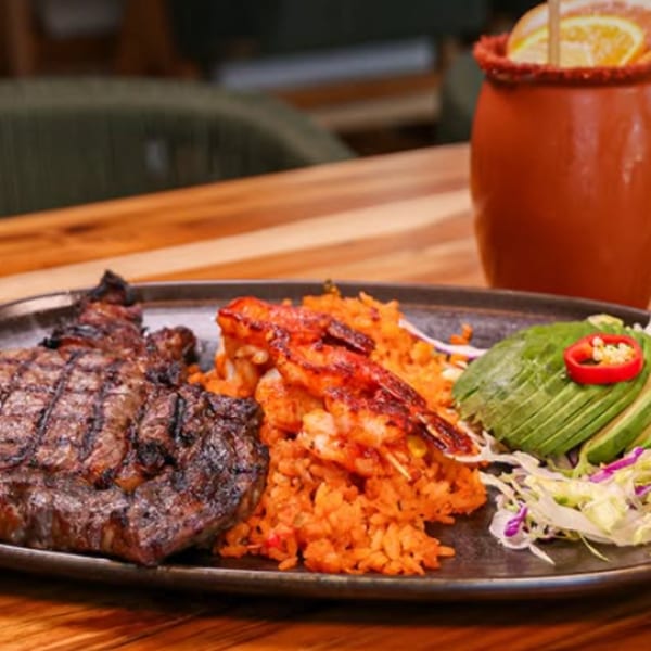 Grilled meat and orange rice served with sliced avocado, salad, and tomato garnish on a dark plate beside a red drink.